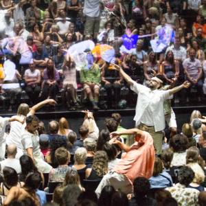 Uns ballarins es mouen per entremig d'una platea de teatre plena de públic. A on hauria de ser l'escenari hi ha un mirall gegant que reflecteix l'escena.