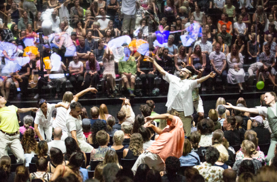 Uns ballarins es mouen per entremig d'una platea de teatre plena de públic. A on hauria de ser l'escenari hi ha un mirall gegant que reflecteix l'escena.