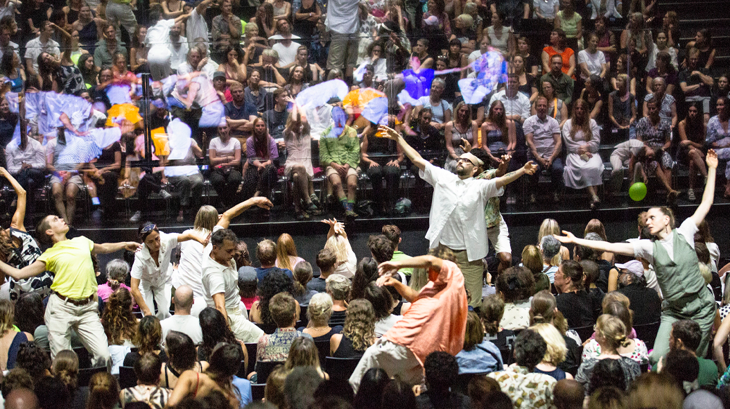 Uns ballarins es mouen per entremig d'una platea de teatre plena de públic. A on hauria de ser l'escenari hi ha un mirall gegant que reflecteix l'escena.